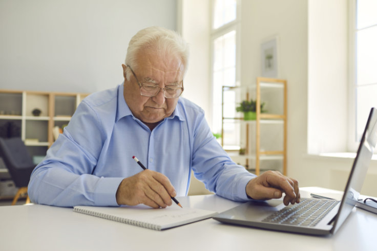 Alter Mann sitzt an einem Laptop und mach sich Notizen